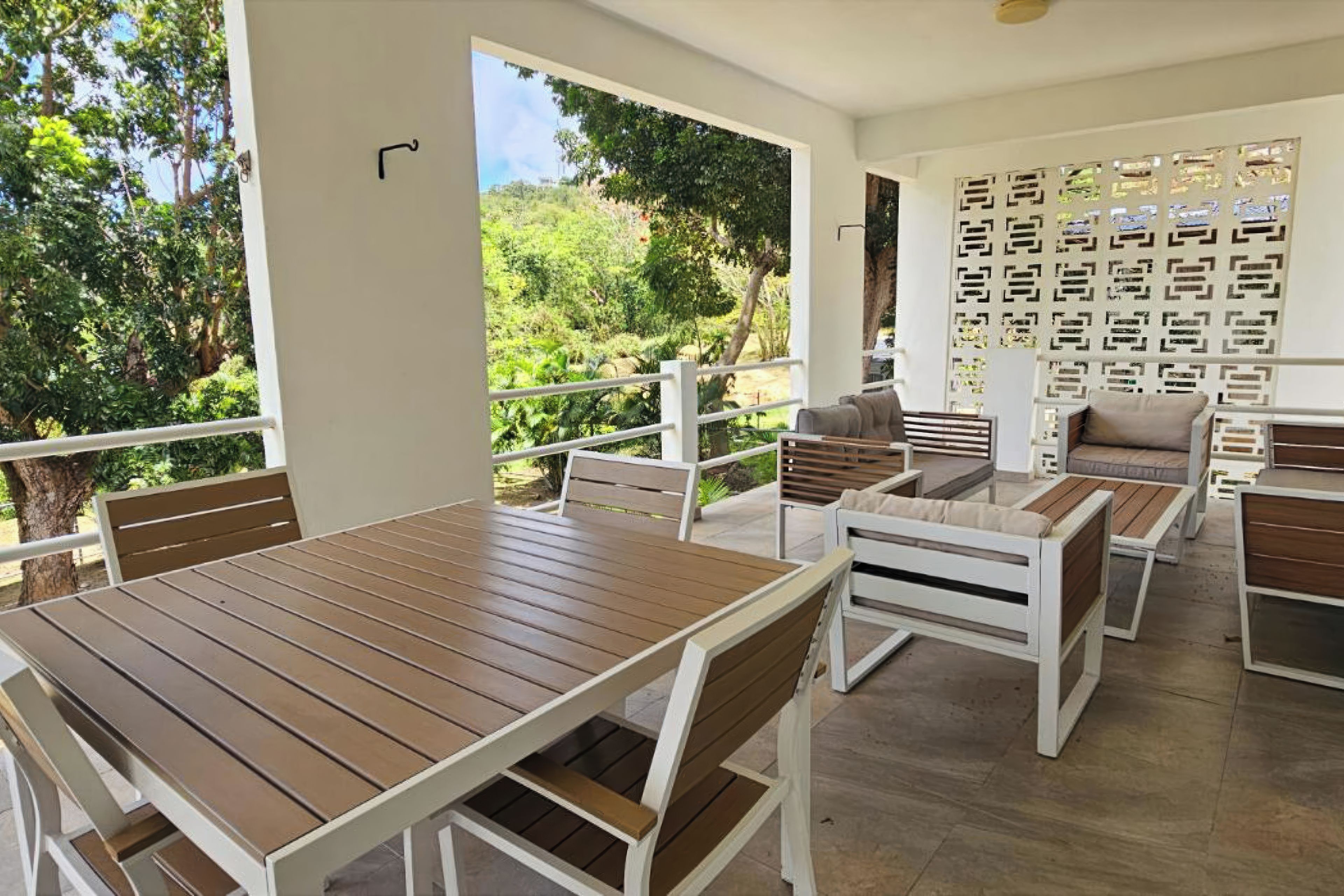 A patio area with a wooden table and benches, a view of greenery, and a white wall with a decorative pattern.