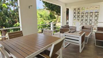 A patio area with a wooden table and benches, a view of greenery, and a white wall with a decorative pattern.