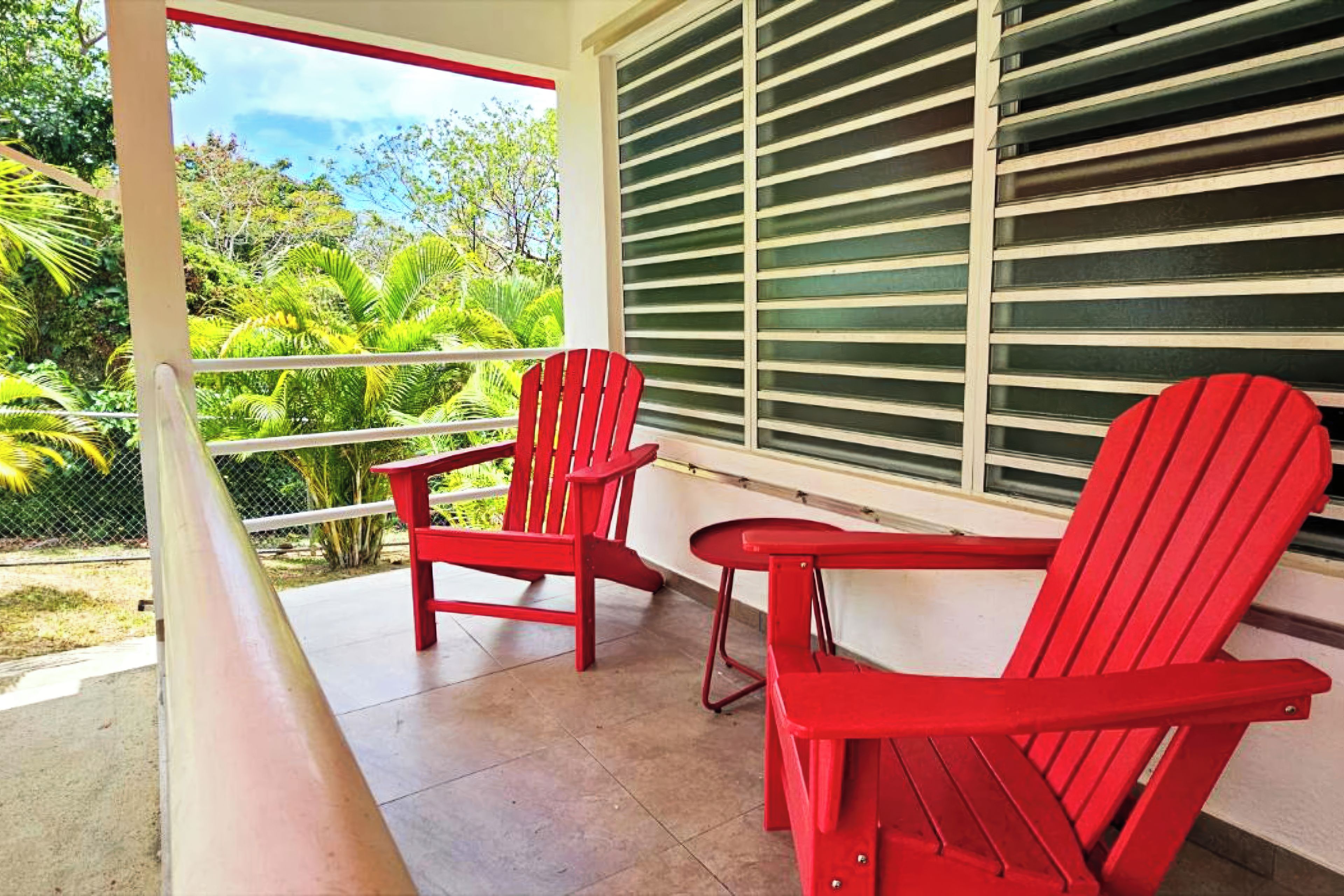A balcony with two red chairs and a table, a window with blinds, and a view of greenery.