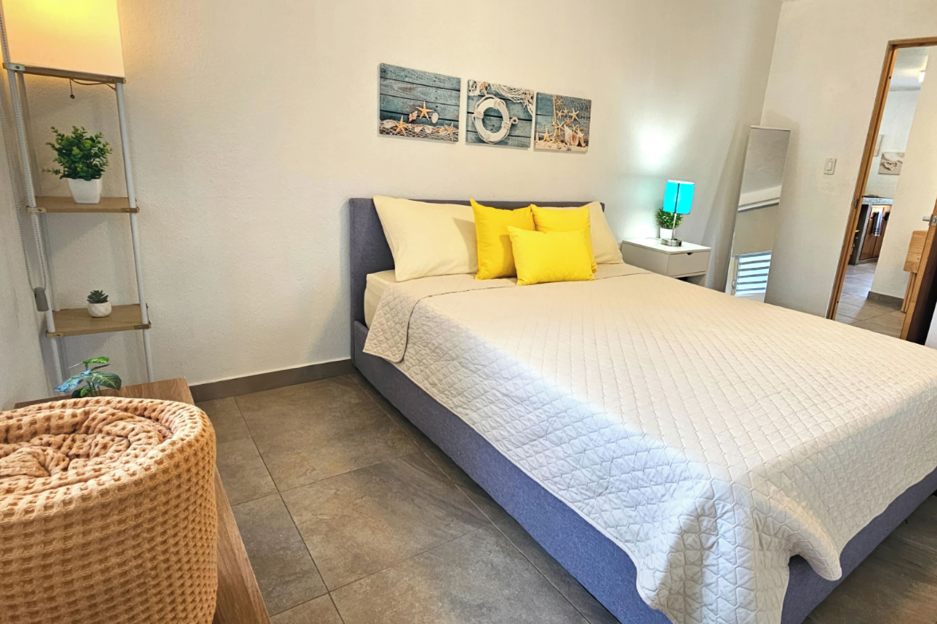 A bedroom with a bed, pillows, a bedside table with a lamp, a wall-mounted shelf with plants, and a door leading to another room.