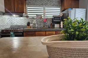 A kitchen with a gas stove, microwave, and stainless steel refrigerator. There is a sign on the window, a potted plant, and wooden cabinets.