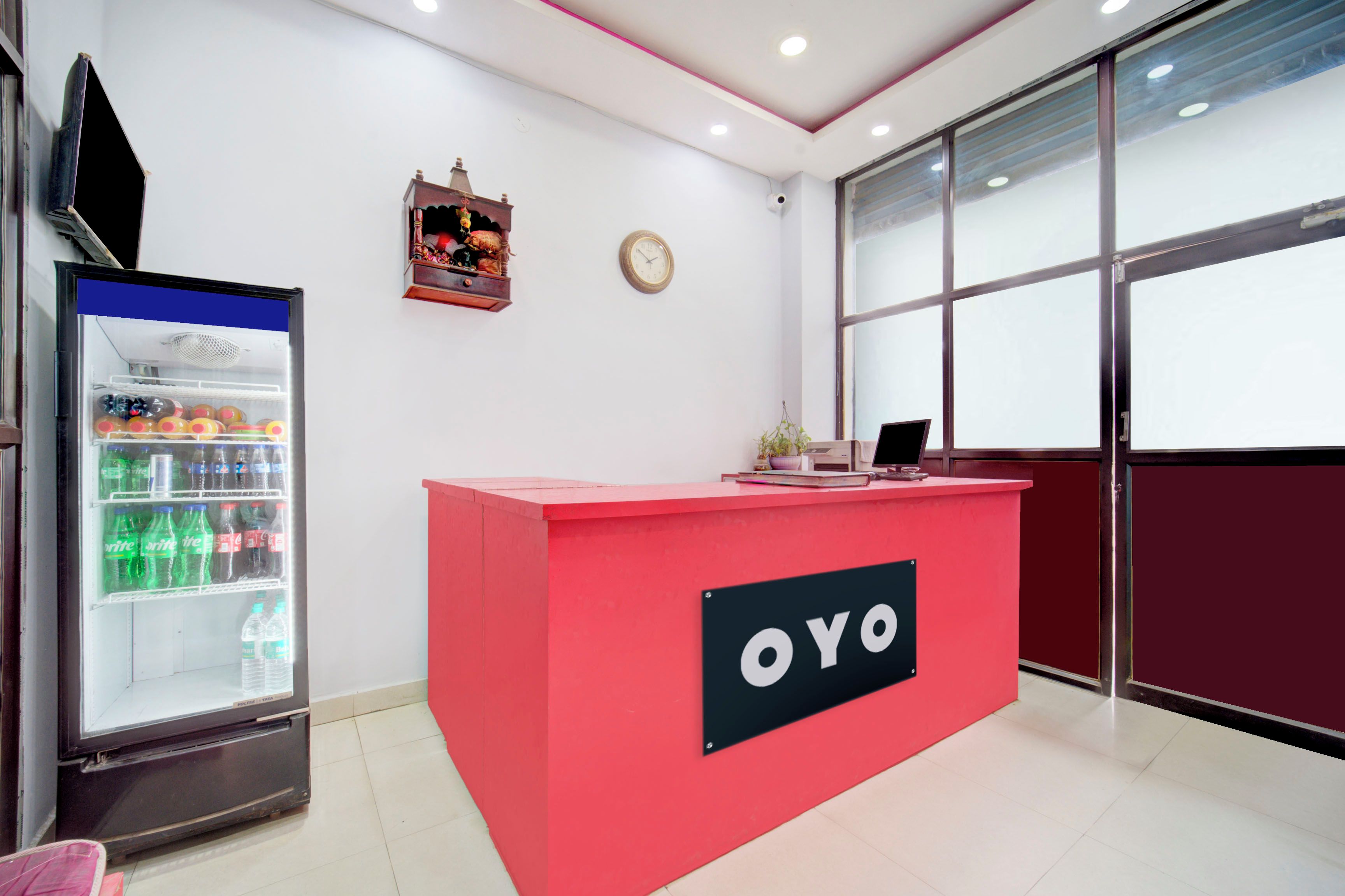 Un mostrador de recepción con un letrero que dice 'OYO' en un espacio de oficina moderno, con un refrigerador lleno de bebidas y un reloj en la pared.