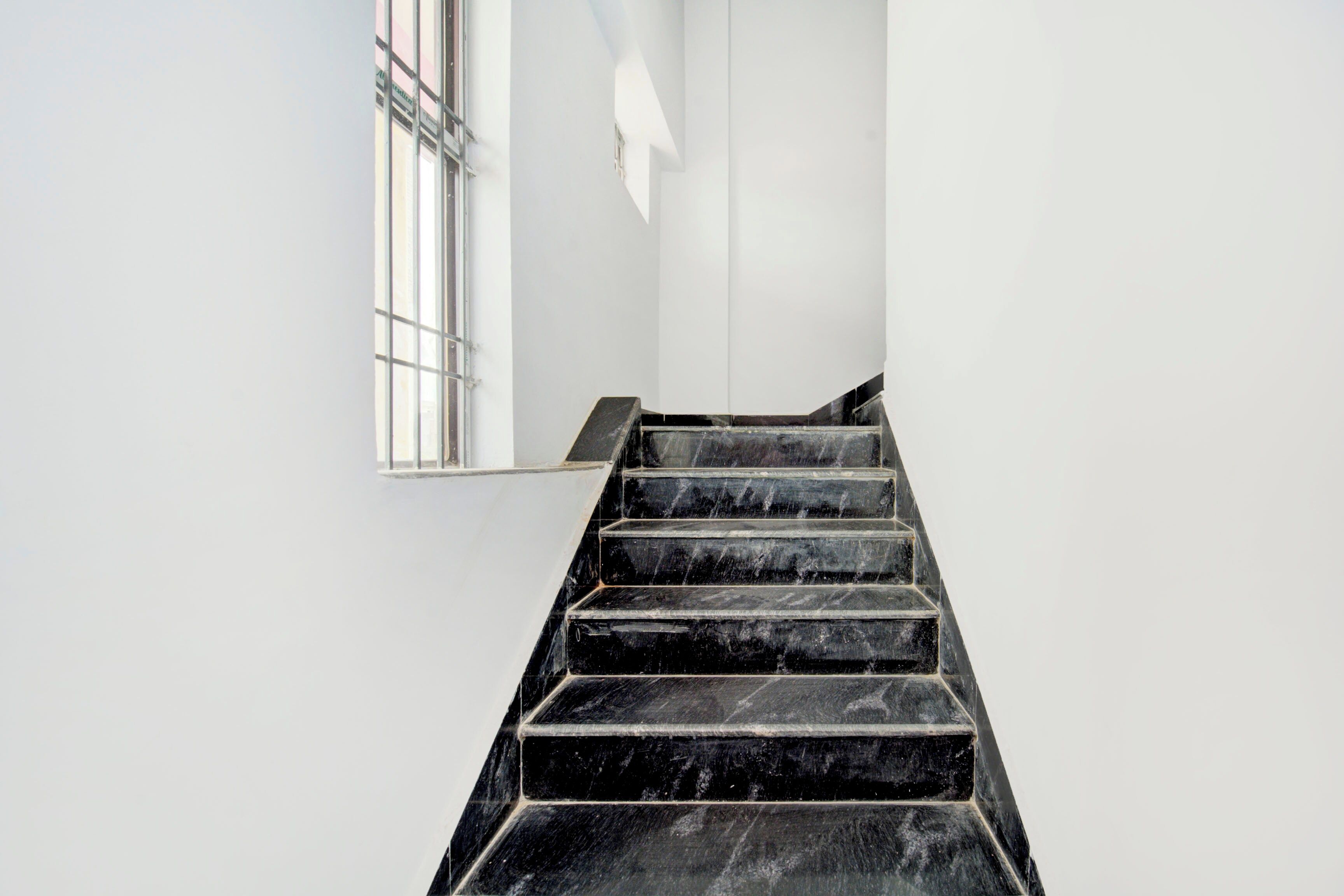 A staircase with black steps leading upwards, white walls, and a window with metal bars.
