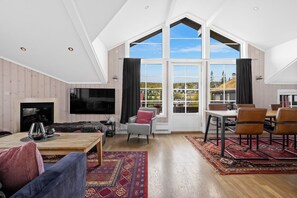 Ein geräumiges Wohnzimmer mit großem Fernseher, Kamin, Essbereich mit Stühlen und Blick durch große Fenster.