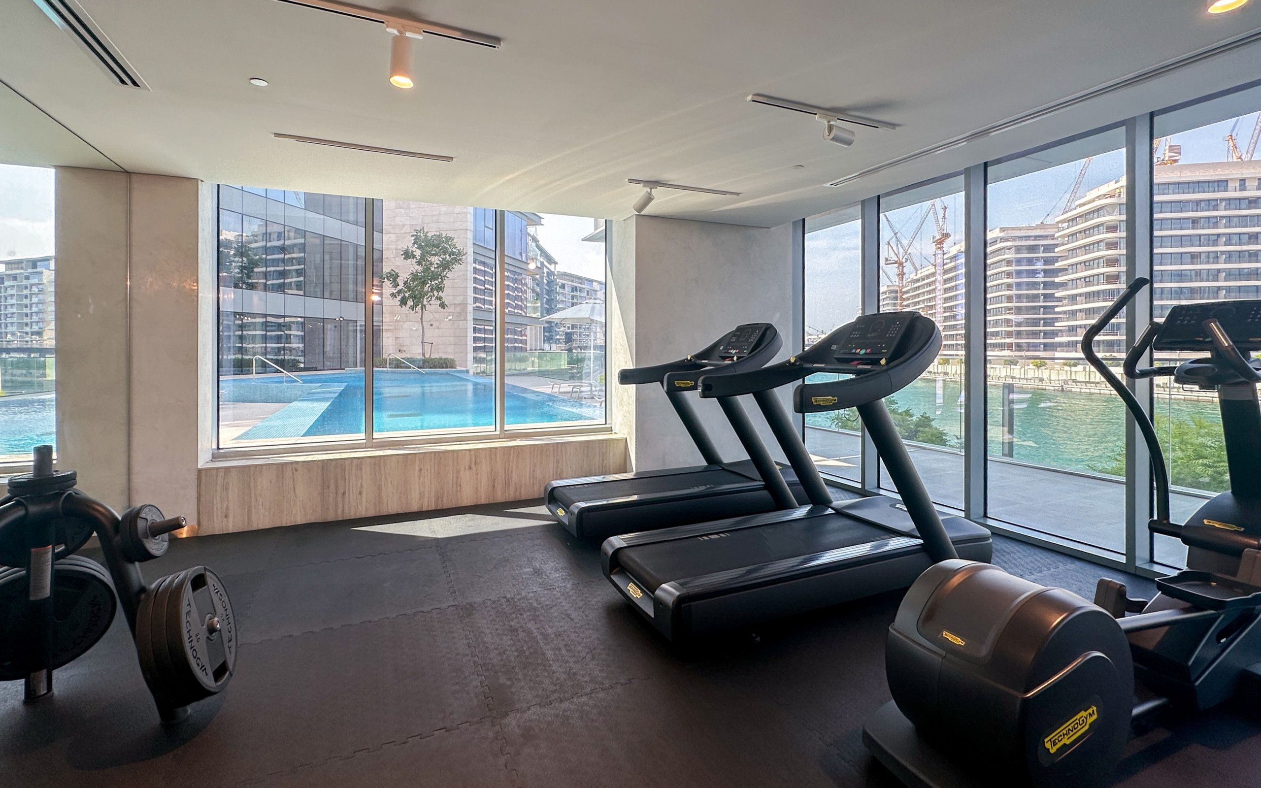 A gym with treadmills, a rowing machine, and a bench press. There is a large window with a view of a swimming pool and buildings.