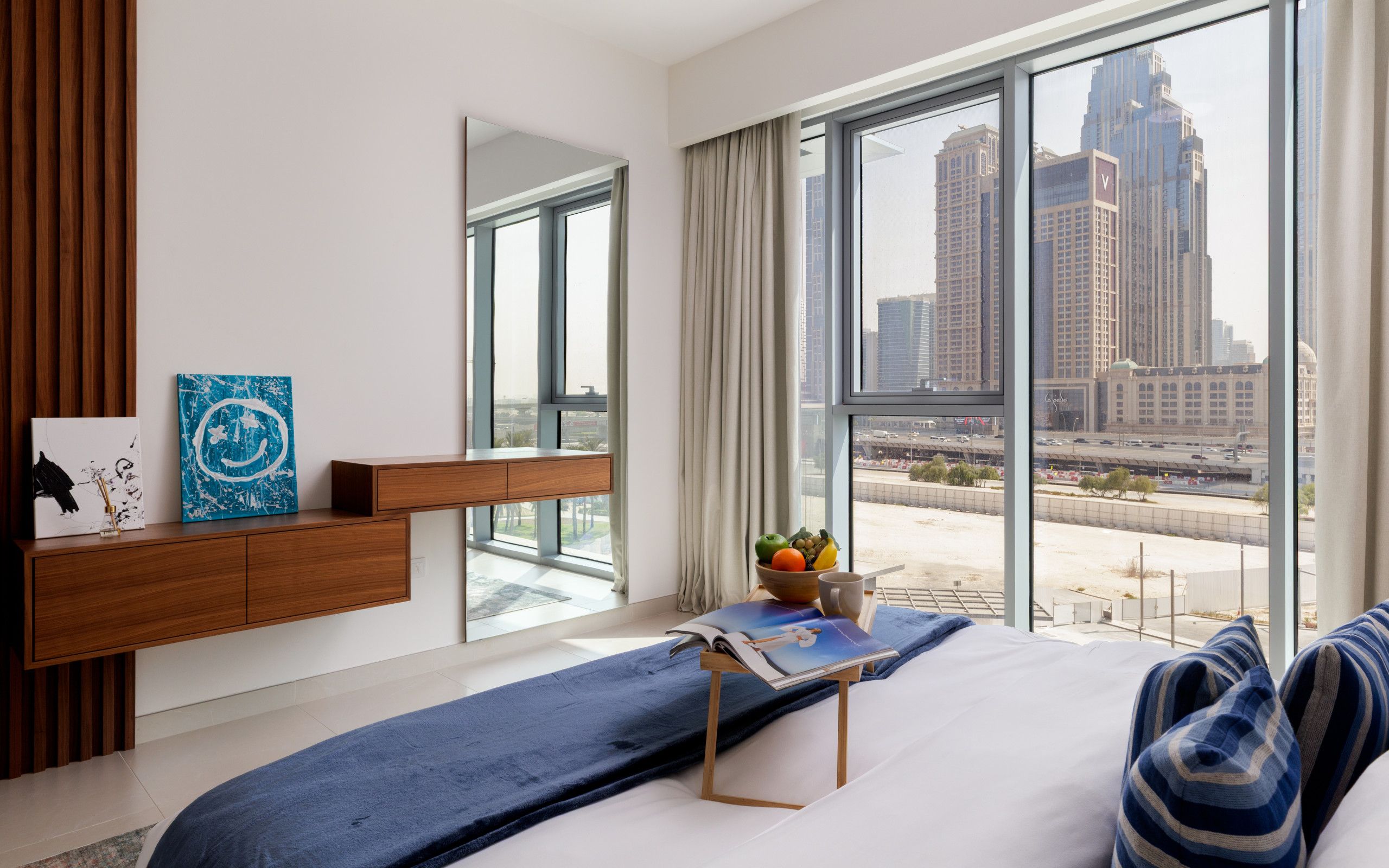A modern hotel room with a large window, a wooden console table, a blue sofa, and a view of a cityscape.