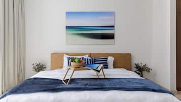 A hotel room with a bed, a bedside table with a bowl of fruit, a painting on the wall, and a view of the sea.