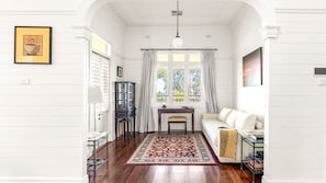 A room with a wooden floor, a sofa, a table with chairs, a cabinet, and a window with curtains. - 1912 Heritage House | Kiama Coastal Living (Kiama)