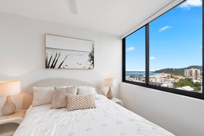 Ein Schlafzimmer mit großem Fenster, einem Bett mit weißer Bettwäsche, zwei Nachttischlampen und einem Wandbild im Strandhausstil.