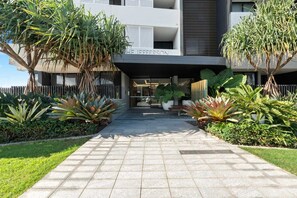Um edifício moderno com entrada pavimentada, rodeado por plantas tropicais e uma cerca de madeira.