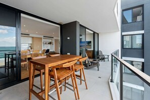 Ein moderner Balkon mit Holztisch und -stühlen, eine Glastür zum Küchenbereich und Blick aufs Meer.