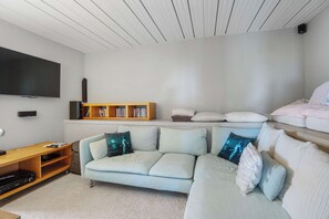 Ein Wohnzimmer mit einem hellen Ecksofa, einem flachbildfernseher an der Wand, einer Holzunterhaltungsecke mit Büchern und einem Bett mit Kissen.