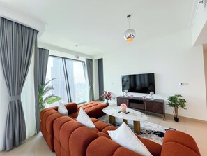 A modern living room with a sectional sofa, a round coffee table, a flat-screen TV, and a large window with curtains. - Stylish 2BR apartment with iconic Burj Khalifa & Dubai Fountain View Downtown (Dubai)