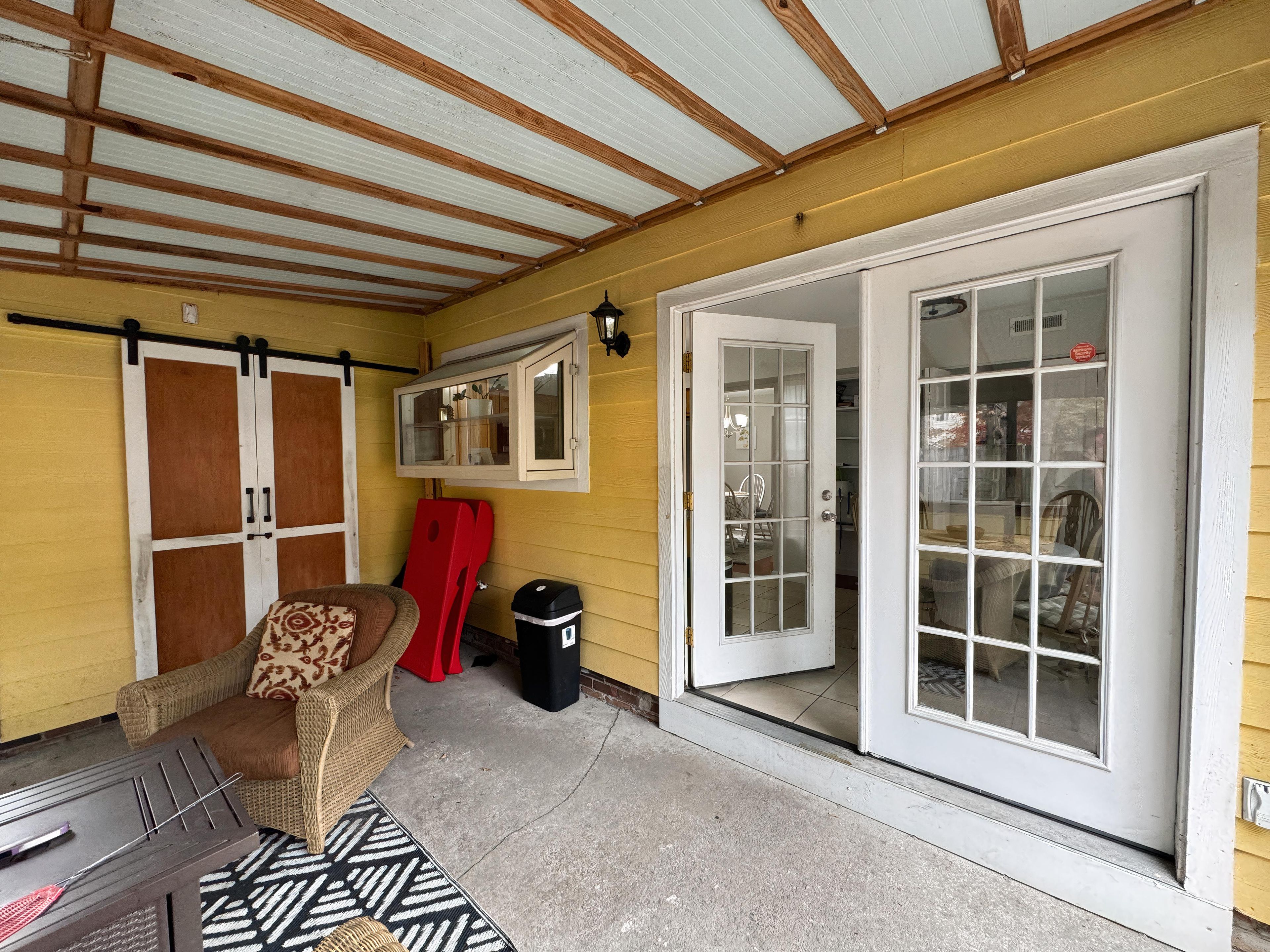 Un espacio exterior cubierto con una puerta corrediza de vidrio que da a un ambiente interior, una silla de mimbre y un cubo de basura.