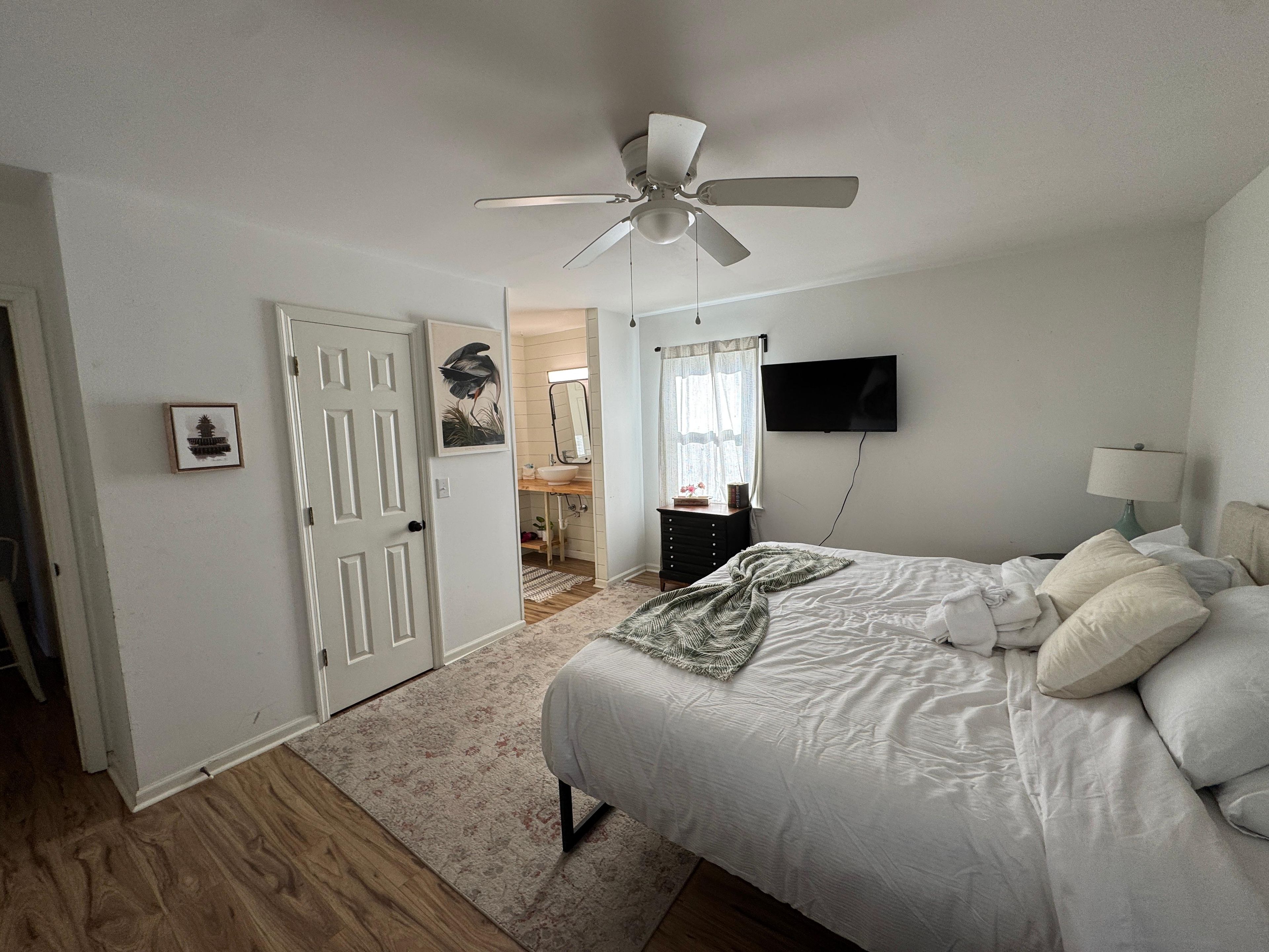 Ein Schlafzimmer mit Bett, Deckenventilator, Fernseher und einem durch eine offene Tür einsehbarer Badezimmer.
