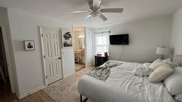 Ein Schlafzimmer mit Bett, Deckenventilator, Fernseher und einem durch eine offene Tür einsehbarer Badezimmer.
