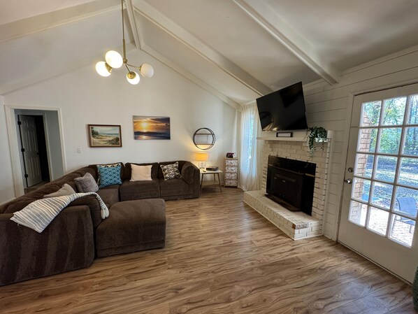 A living room with a sectional sofa, a fireplace, a flat-screen TV, and a large window. - Plush in the Pines! - 4BR + Game Room (Ladson)