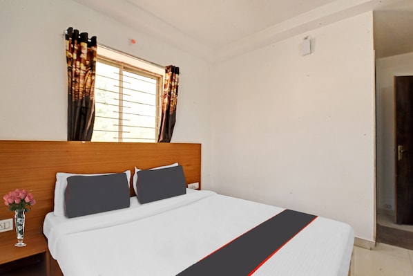 A hotel room with a wooden headboard, a bed with white bedding and a black and red accent, a window with patterned curtains, and a vase with pink flowers on the nightstand.