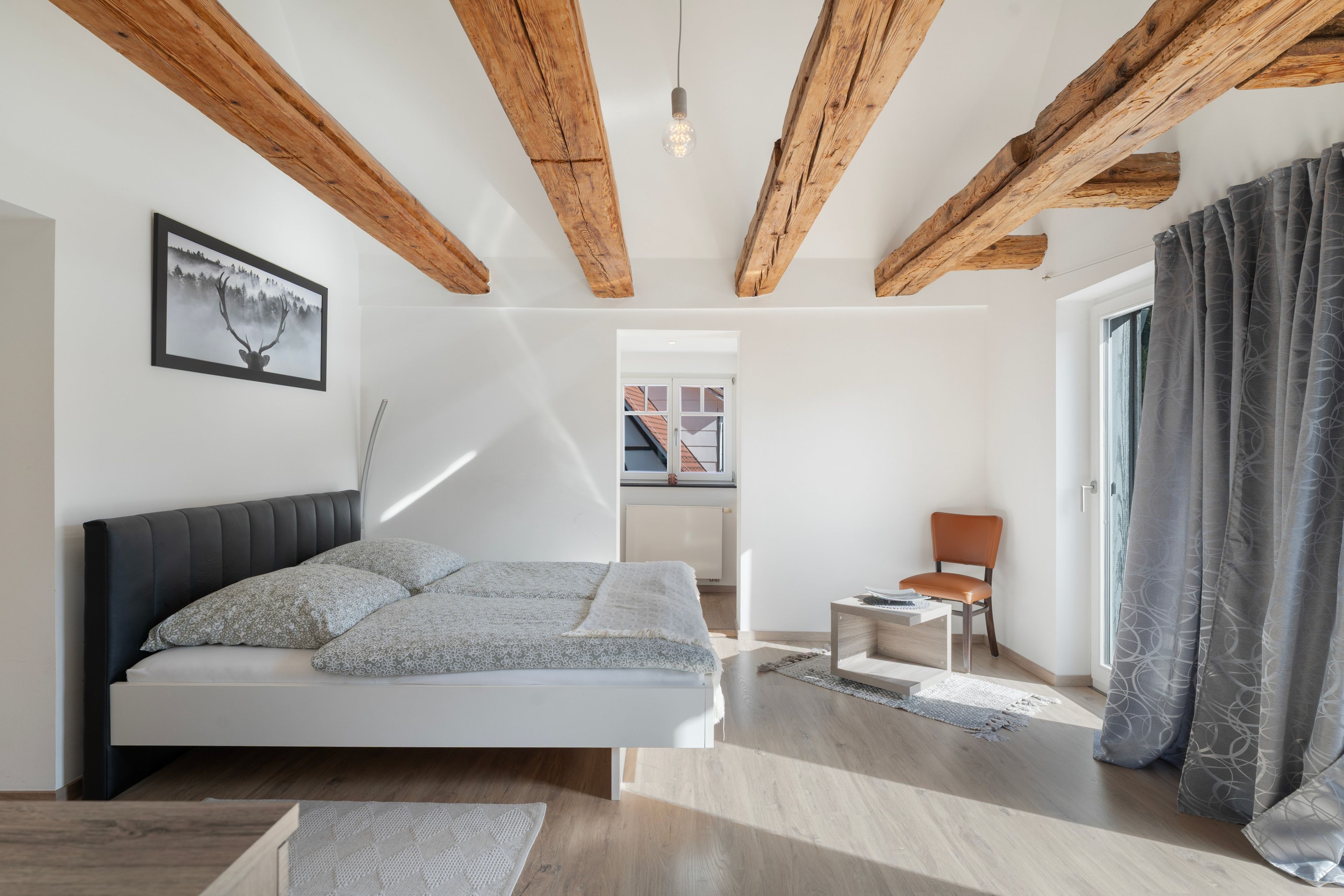 Een moderne slaapkamer met een groot bed, houten balken, een ingelijste foto en uitzicht naar buiten.
