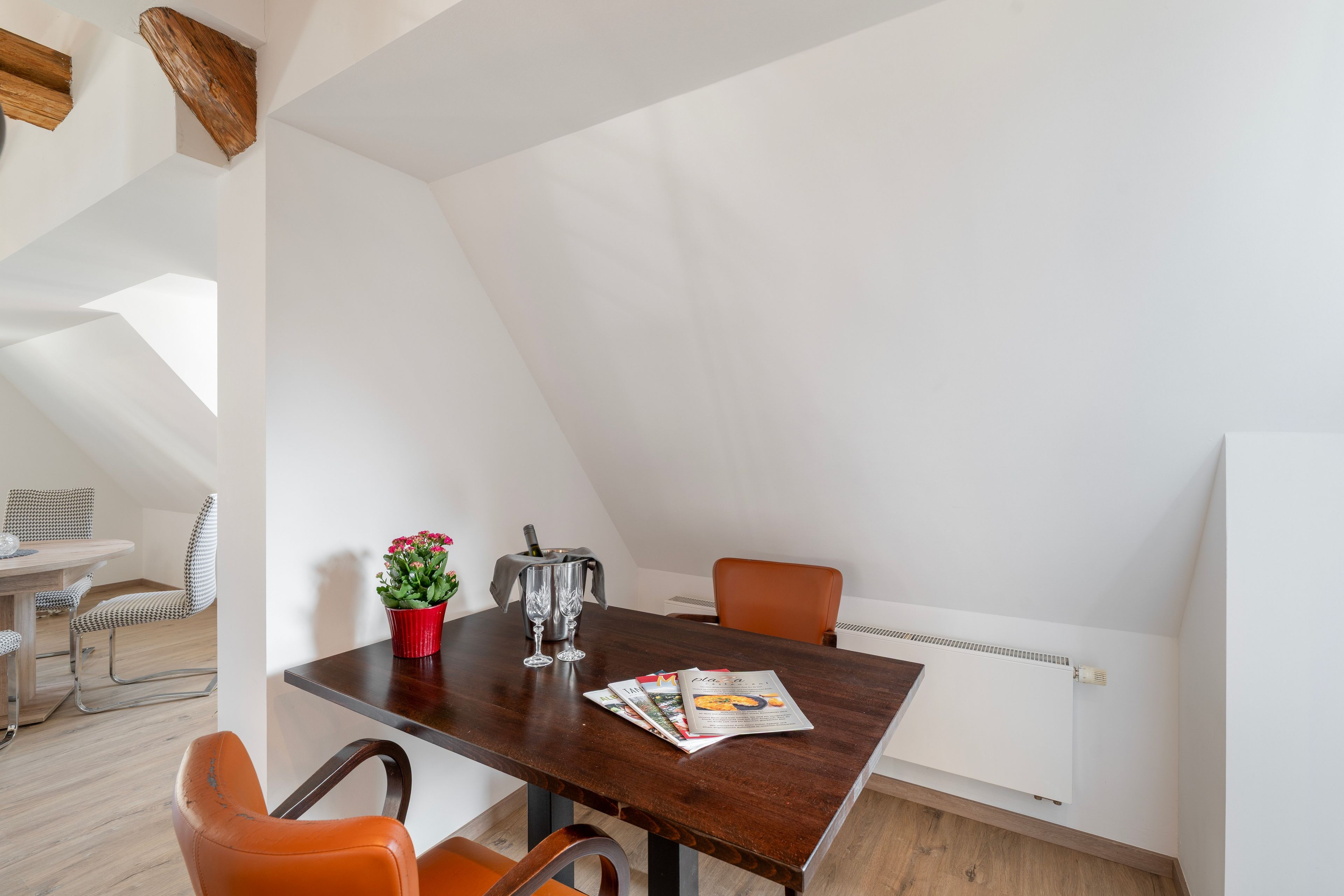 Un coin repas avec une table en bois, deux chaises, un pot de fleurs rouge et un magazine.