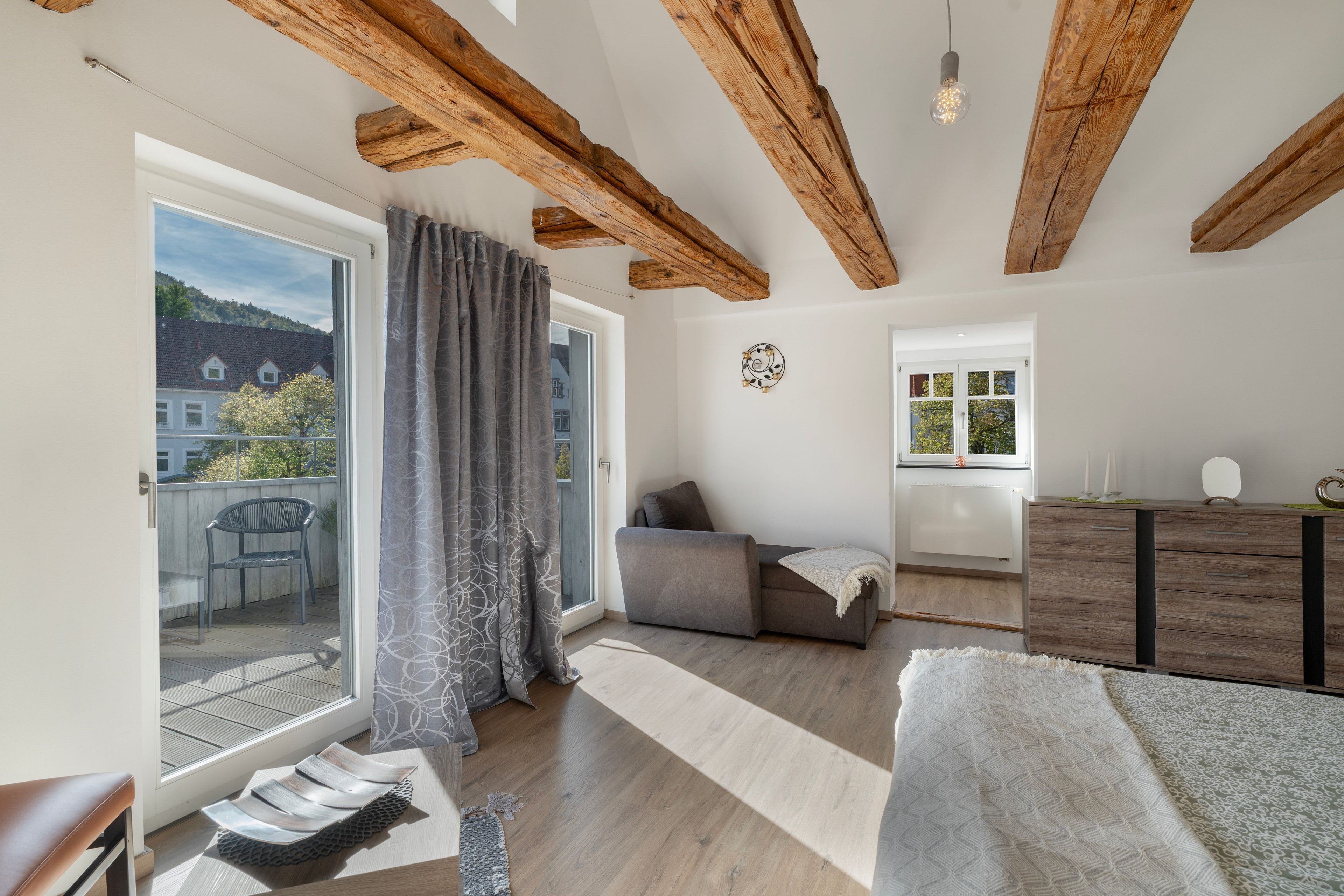 Ein modernes Zimmer mit Balkon, Holzbalken, einer Couch, einem Schreibtisch und Blick auf Gebäude.