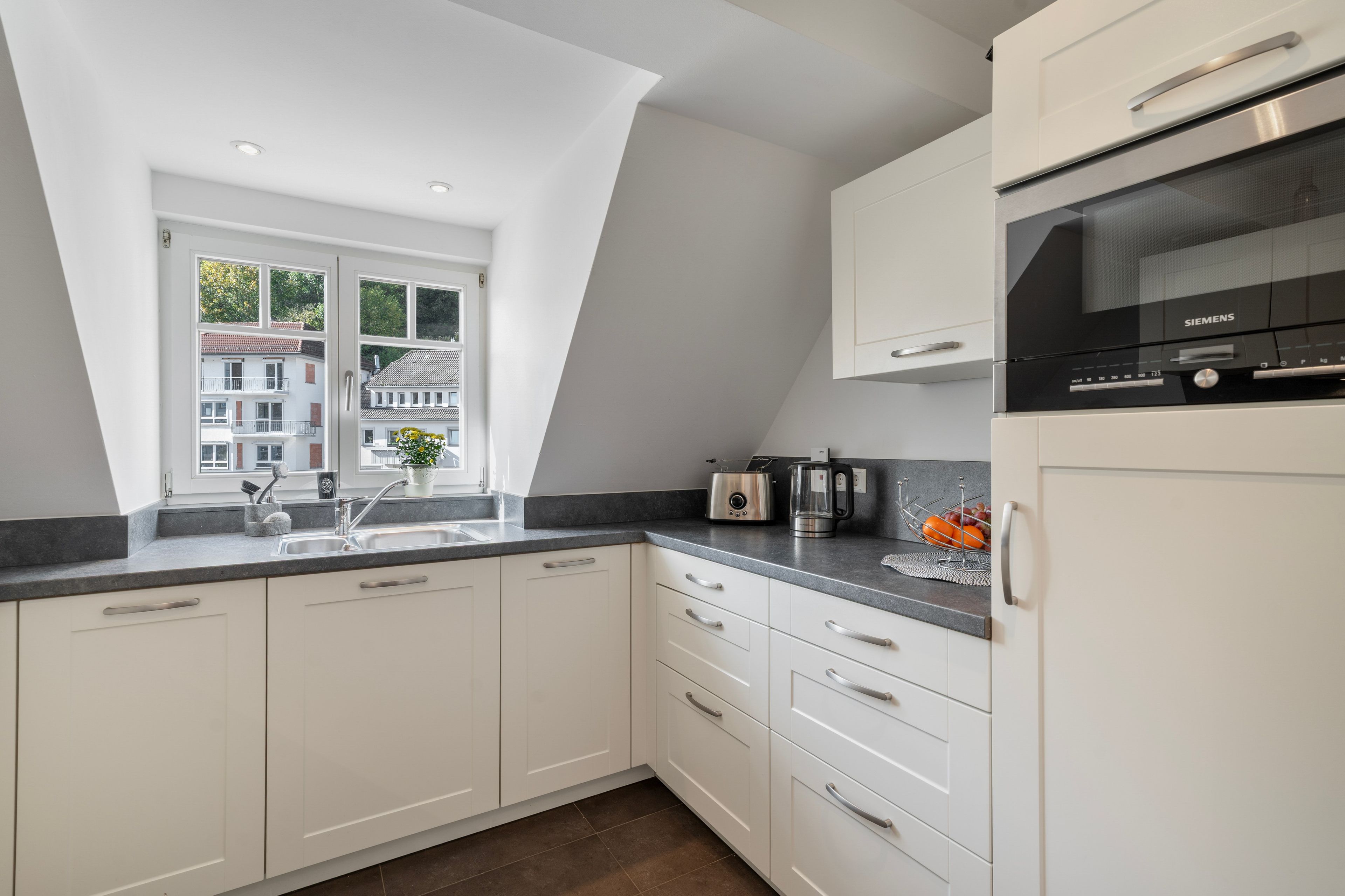 Une cuisine moderne avec des meubles blancs, un four encastré et une fenêtre donnant sur un quartier résidentiel.