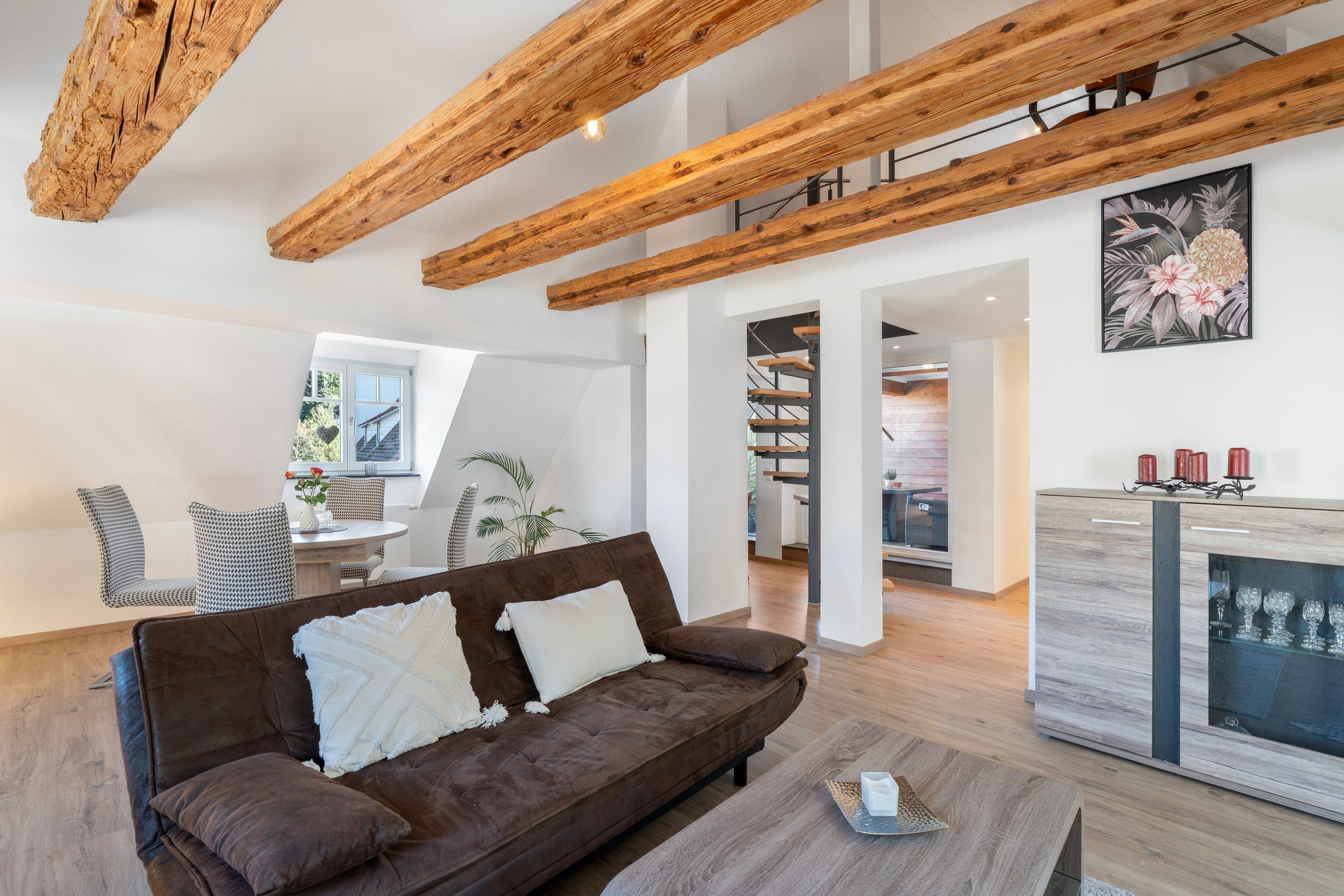 Ein modernes Wohnzimmer mit einem braunen Sofa, Holzbalken, einem Essbereich mit gemusterten Stühlen und einer Küche mit einer Holzarbeitsplatte.