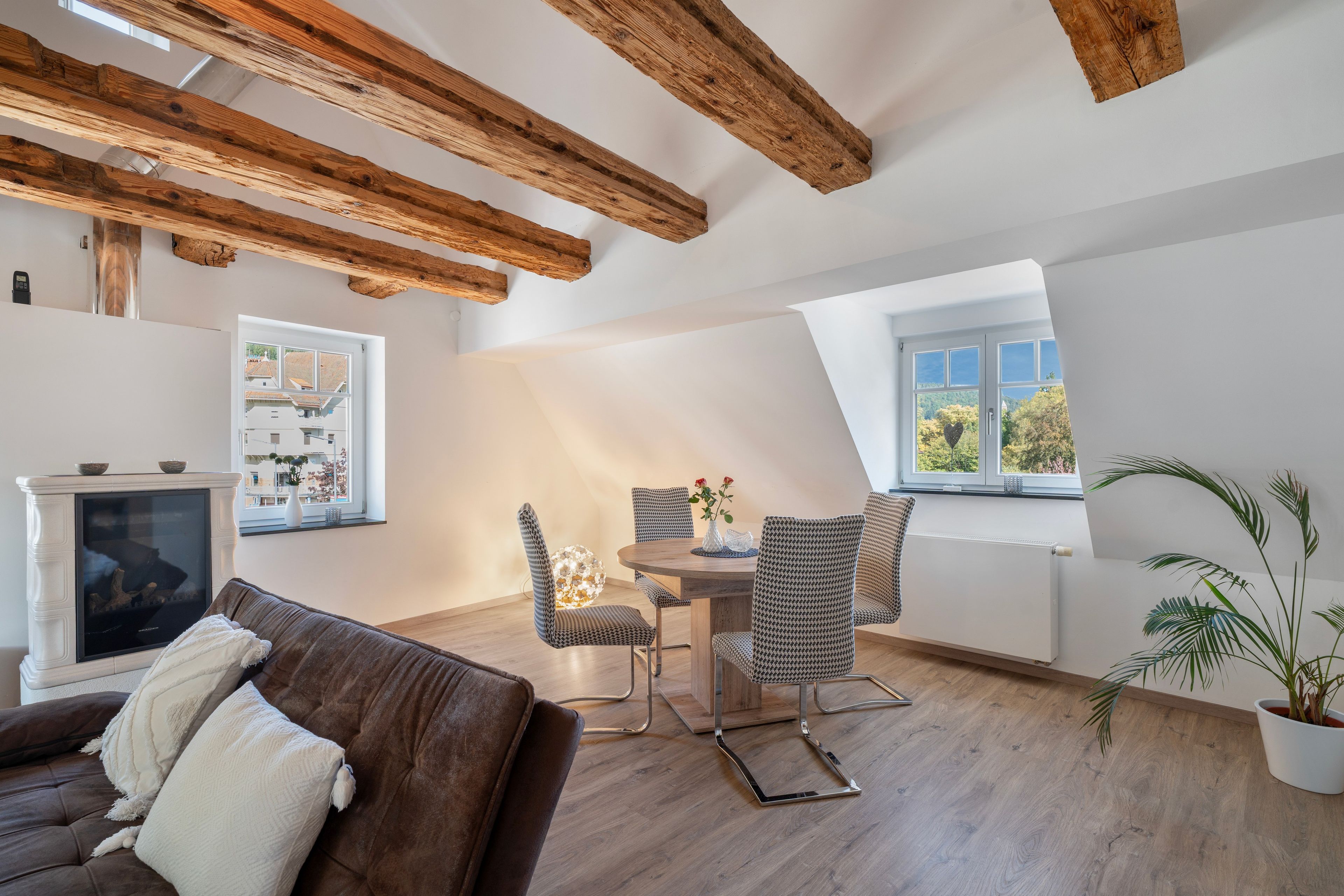 Een moderne woonkamer met zichtbare houten balken, een open haard, een eethoek met stoelen en een uitzicht door een raam.