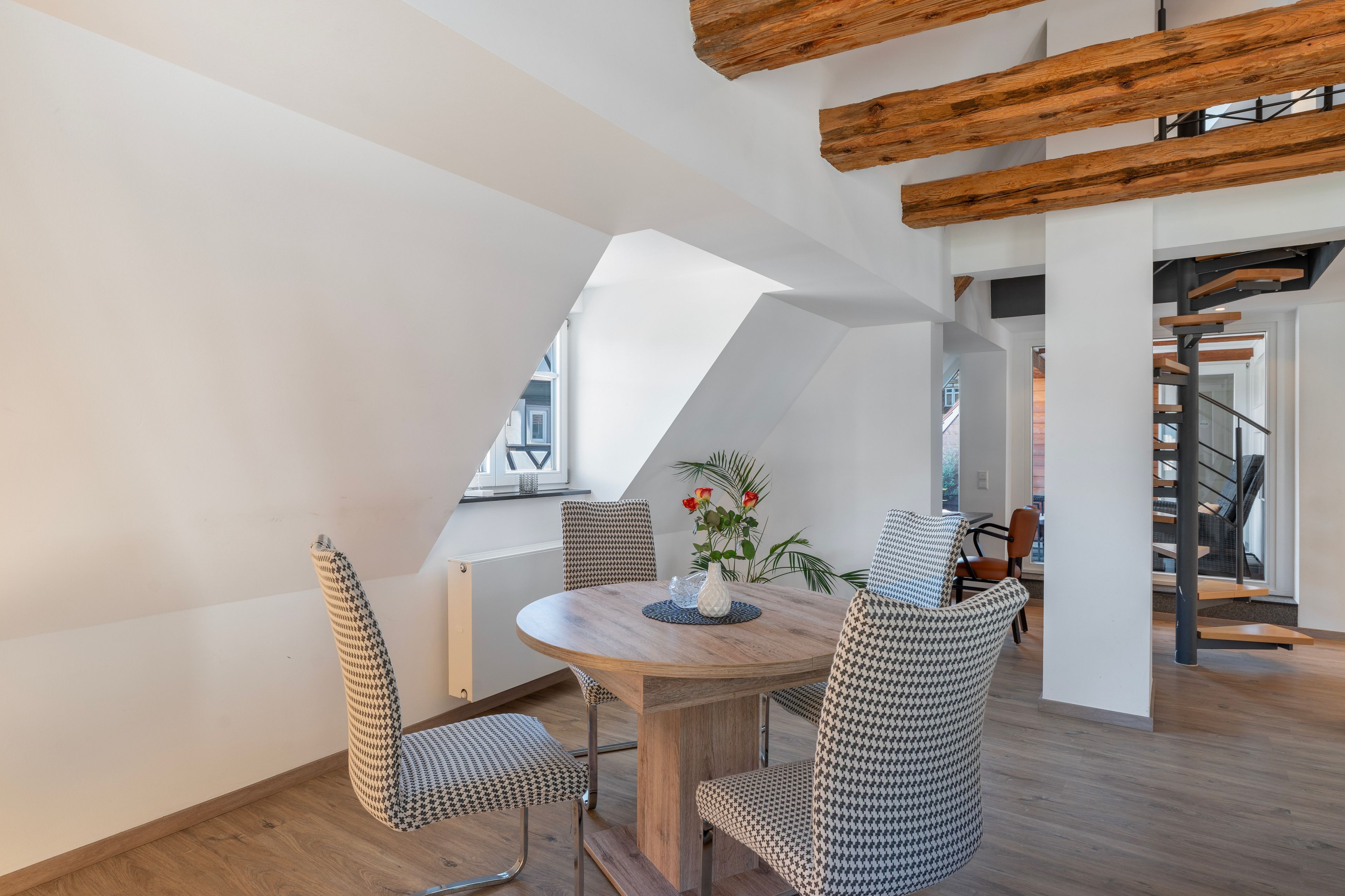 Un coin repas avec une table ronde, des chaises à motifs pied-de-poule, un vase de fleurs et un plafond en bois.