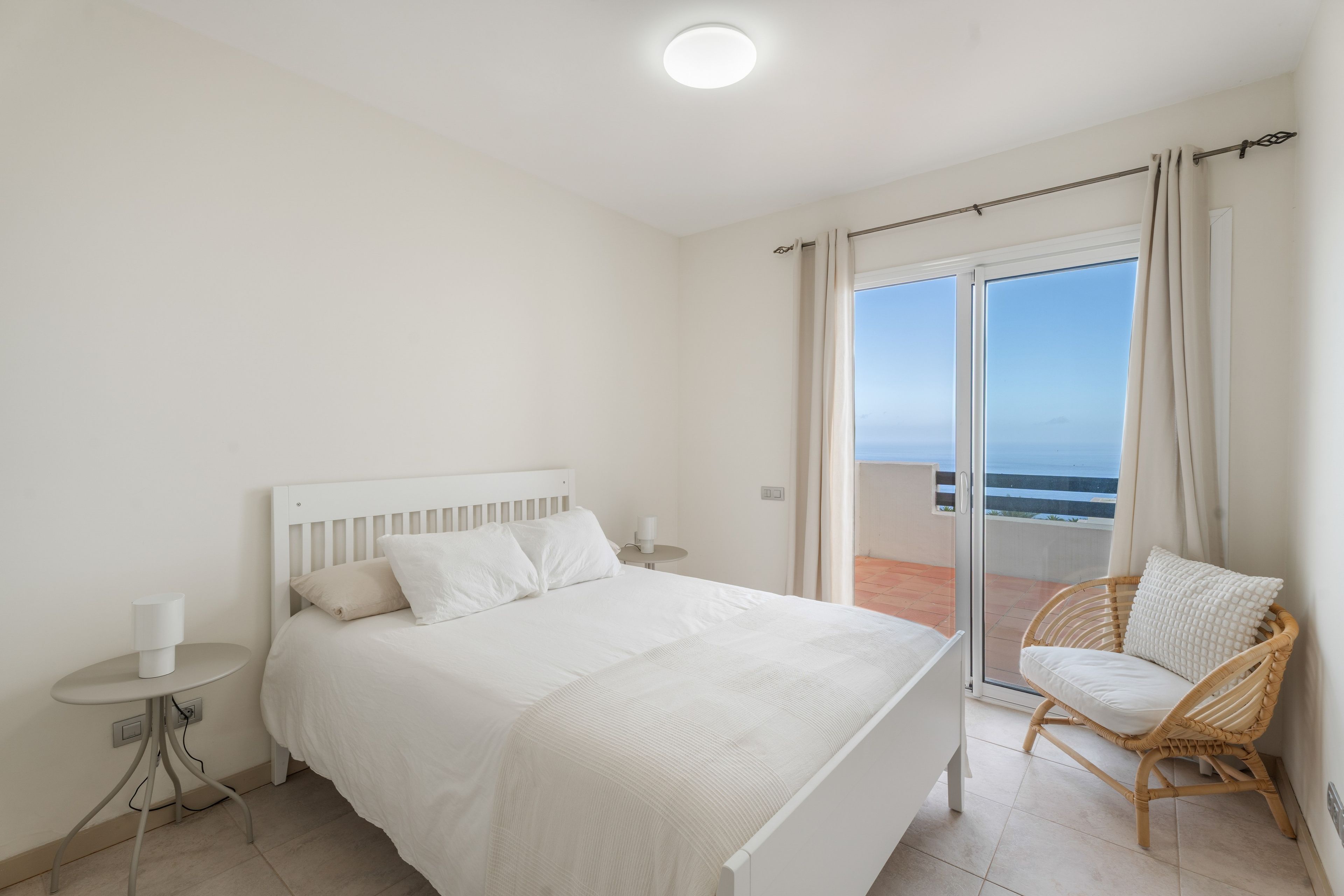 Un dormitorio con cama, mesita de noche, una silla y vistas al mar a través de un ventanal corredero.