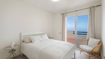 Un dormitorio con cama, mesita de noche, una silla y vistas al mar a través de un ventanal corredero.