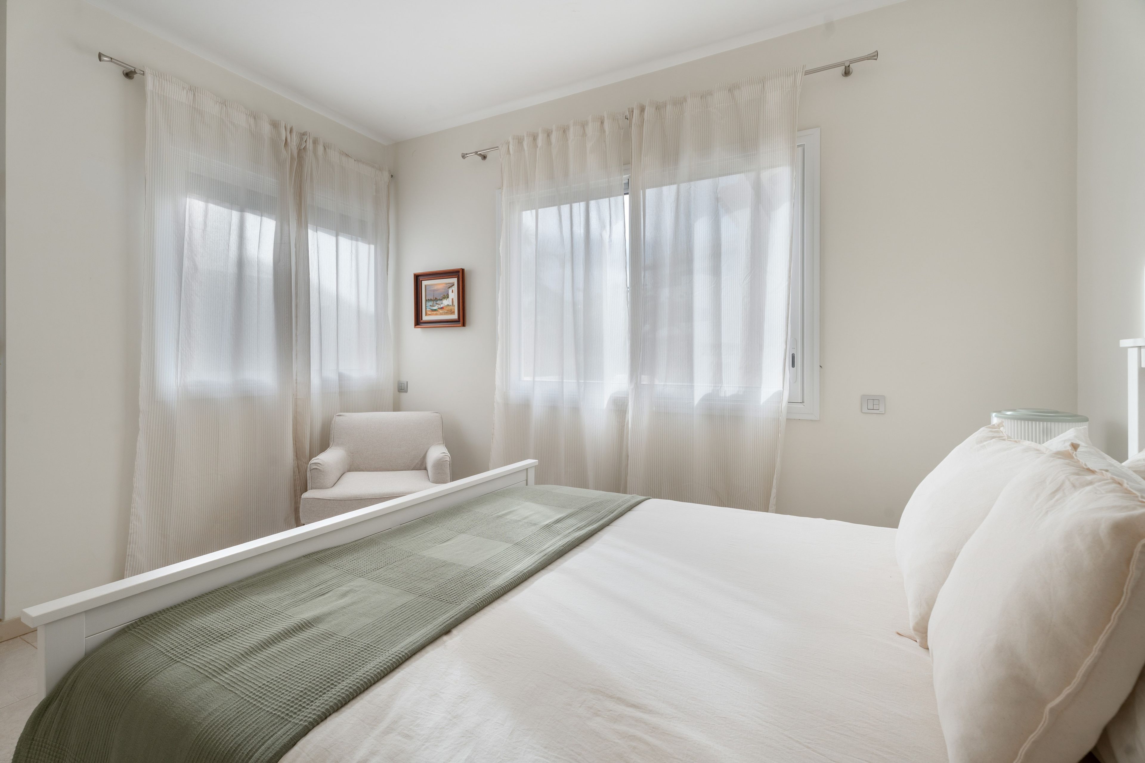Un dormitorio con cama, una silla y dos ventanas con cortinas.