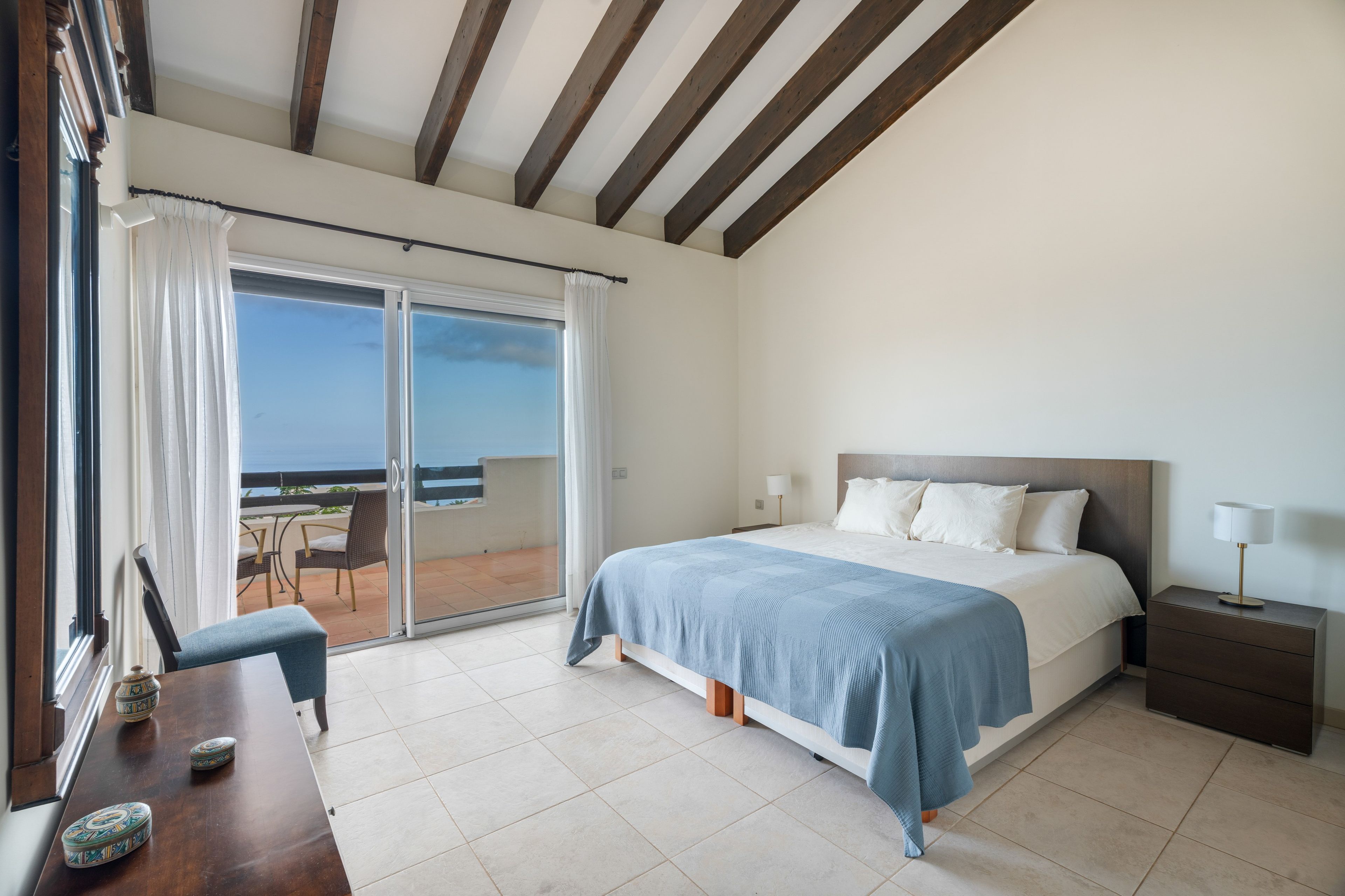 A bedroom with a bed, a nightstand, a table with chairs, and a view of the sea through a sliding door.
