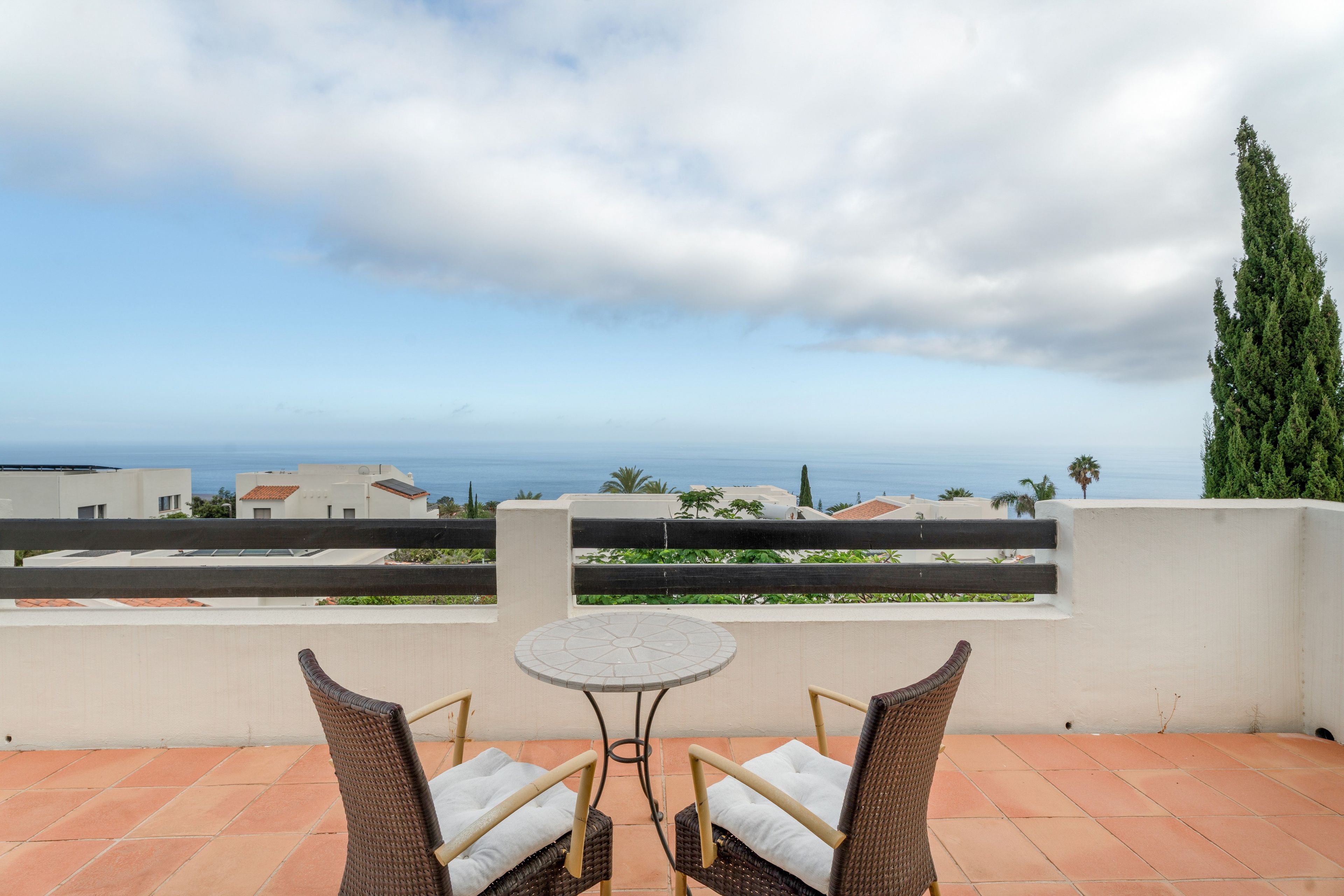 Un balcon avec deux fauteuils en osier et une petite table, offrant une vue sur la côte, avec des bâtiments et un palmier.