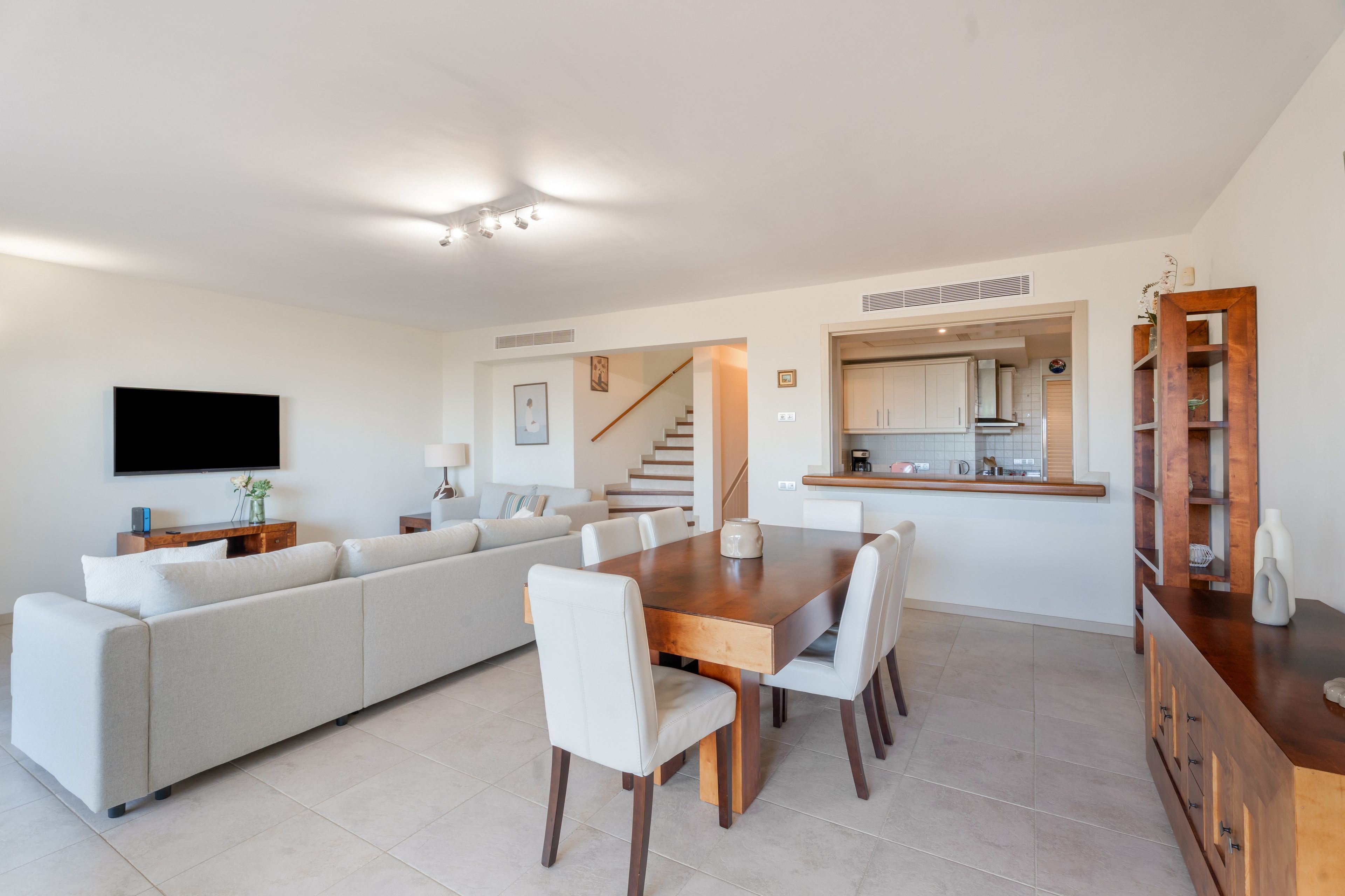 A modern living room with a sectional sofa, dining table, and chairs, a flat-screen TV, and a kitchen area with cabinets and a wooden cabinet.