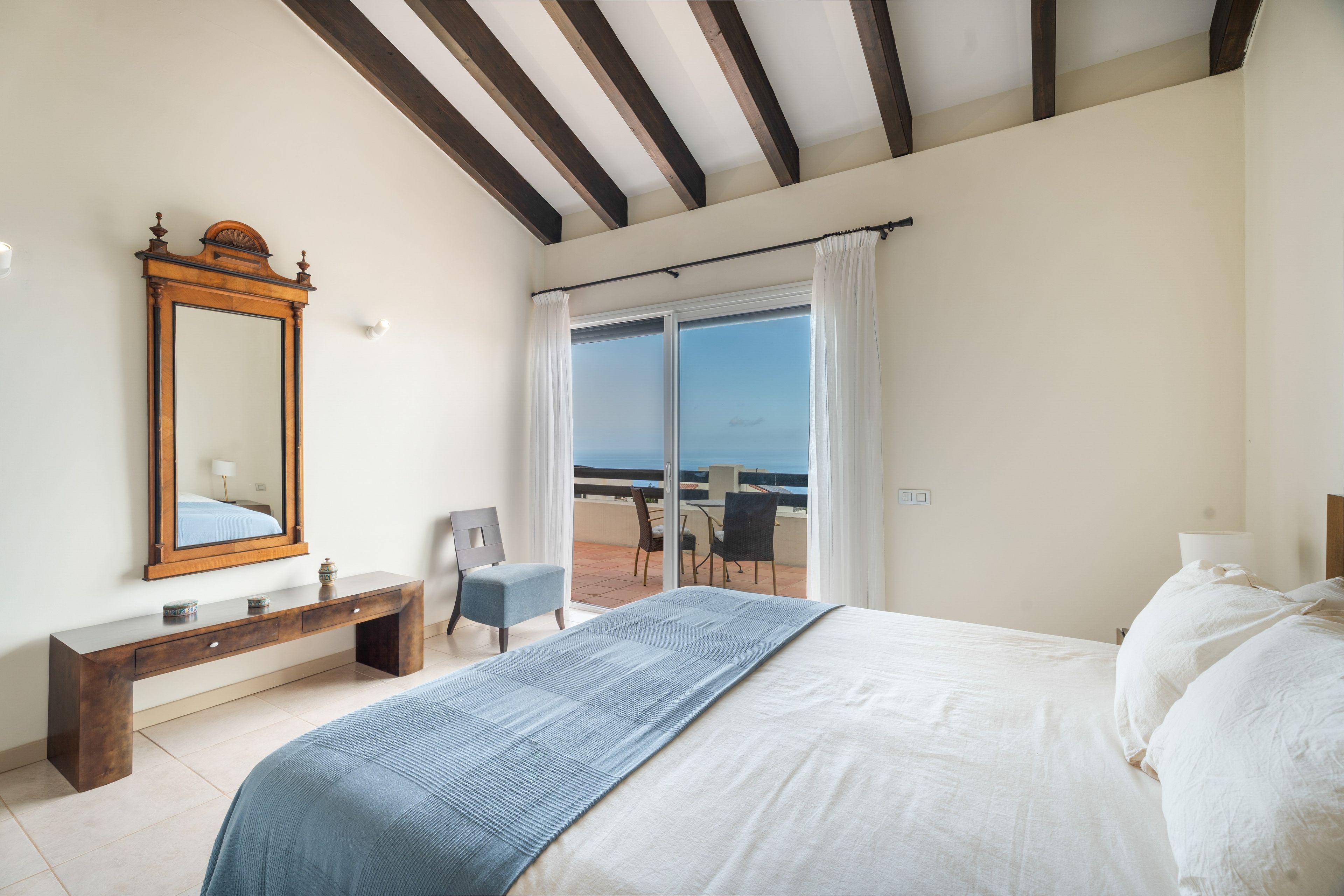 Ein Schlafzimmer mit Bett, Kommode, Spiegel, Stuhl und Blick ins Freie durch eine Glastür.