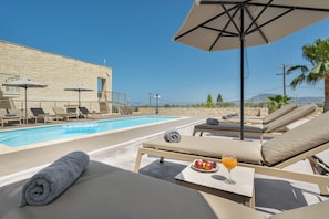 Ein Poolbereich mit Liegestühlen, einem Tisch mit Obst und Getränken und Blick auf das Meer und die Berge.
