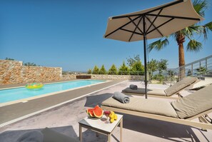 Ein Poolbereich mit Liegestühlen, einem Tisch mit Obst und Getränken und Blick auf das Meer.