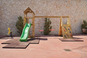 Ein Spielplatz mit einer Holzerne Rutsche, Schaukeln und Klettergerüsten vor einer Steinmauer.