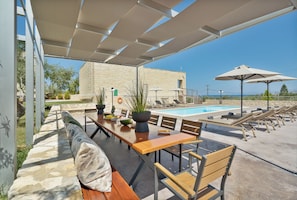 Un espace extérieur avec une piscine, une table et des chaises en bois, un bâtiment en pierre en arrière-plan et un ciel bleu dégagé.
