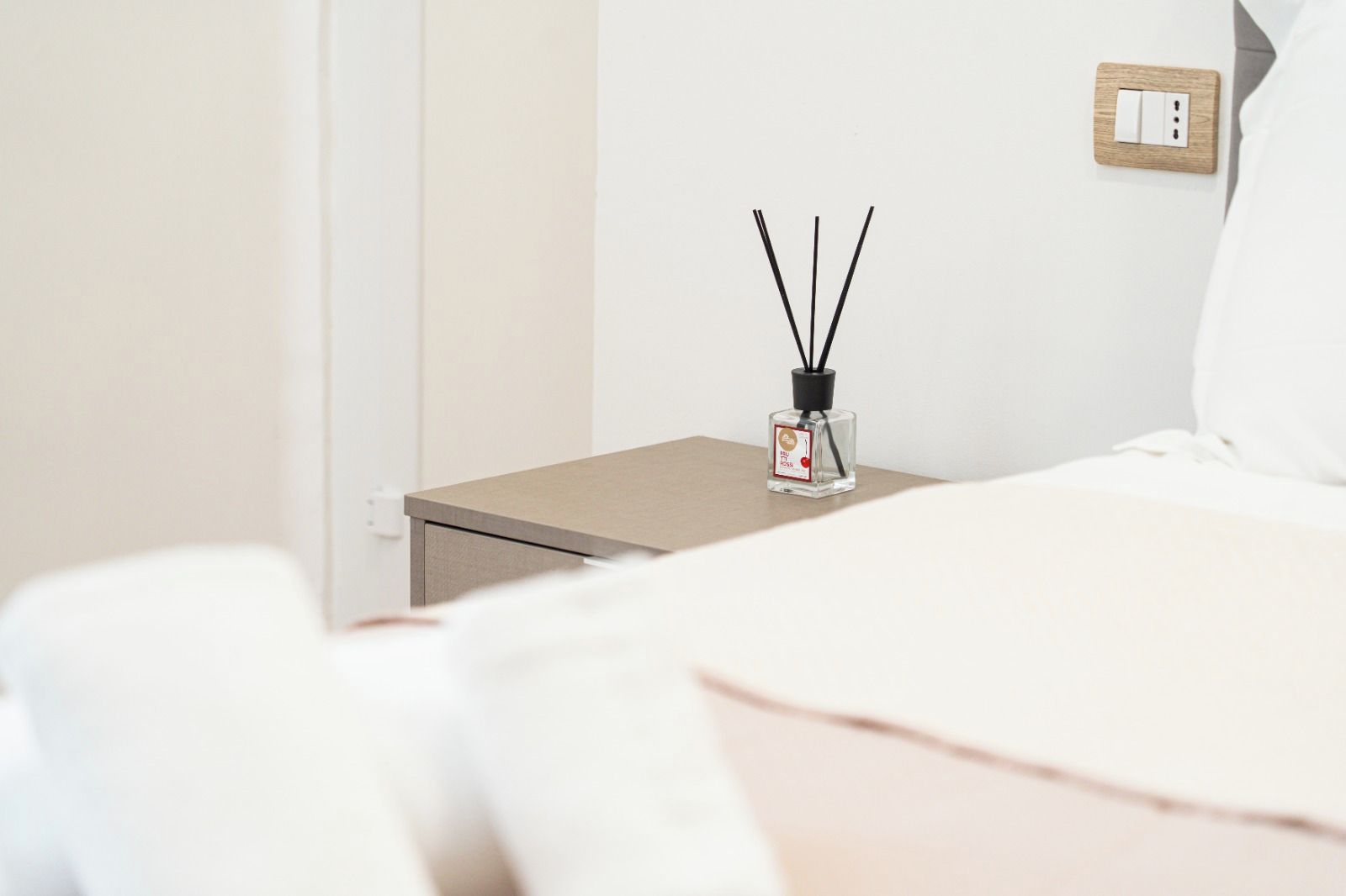 Ein ordentlich bezogenes Bett mit weißen Leinen, ein Nachttisch mit einer kleinen Flasche ätherischem Öl und eine Steckdose an der Wand.