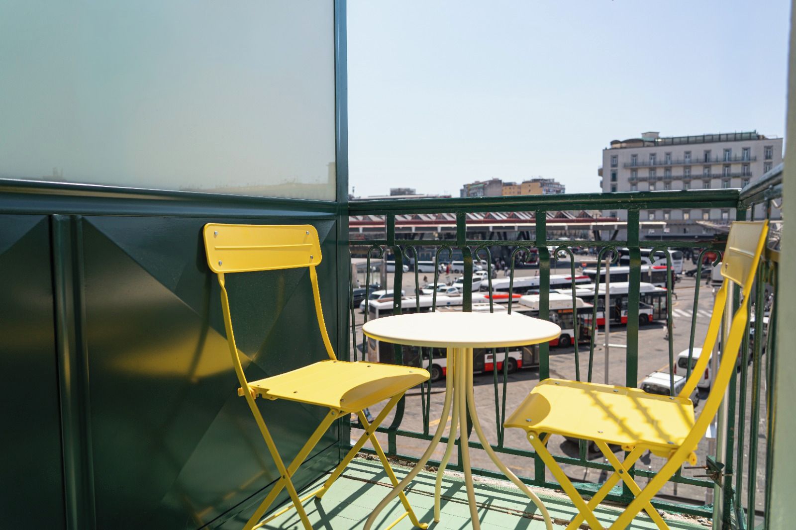Ein Balkon mit gelben Stühlen und einem kleinen runden Tisch, mit Blick auf einen Parkplatz mit Bussen und Gebäuden im Hintergrund.