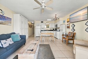 A spacious living room with a blue sofa, a wooden coffee table, and a dining area with a kitchen in the background. - Beachfront condo w Gulf views, 3 pools, tennis & fitness room ~ Sleep 6 (Orange Beach)