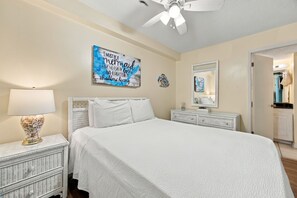 Quarto com cama, mesa de cabeceira, abajur, ventilador de teto e uma frase motivacional na parede.