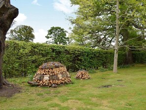 A stack of firewood in a grassy outdoor area with trees and a hedge in the background. - Glebe Cottage | 2 Bedroom Cottage in Perthshire (Scone)