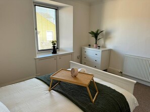 A bedroom with a bed, bedside table, dresser, window, radiator, and a potted plant. - Town Centre Flat For Two Adults (Perth and Kinross)