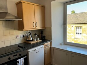 A kitchen with wooden cabinets, a stove, a dishwasher, and a window with a view of a building. - Town Centre Flat For Two Adults (Perth and Kinross)
