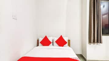 Una cama con ropa blanca y detalles rojos, una ventana con cortinas y un interruptor de luz en la pared.