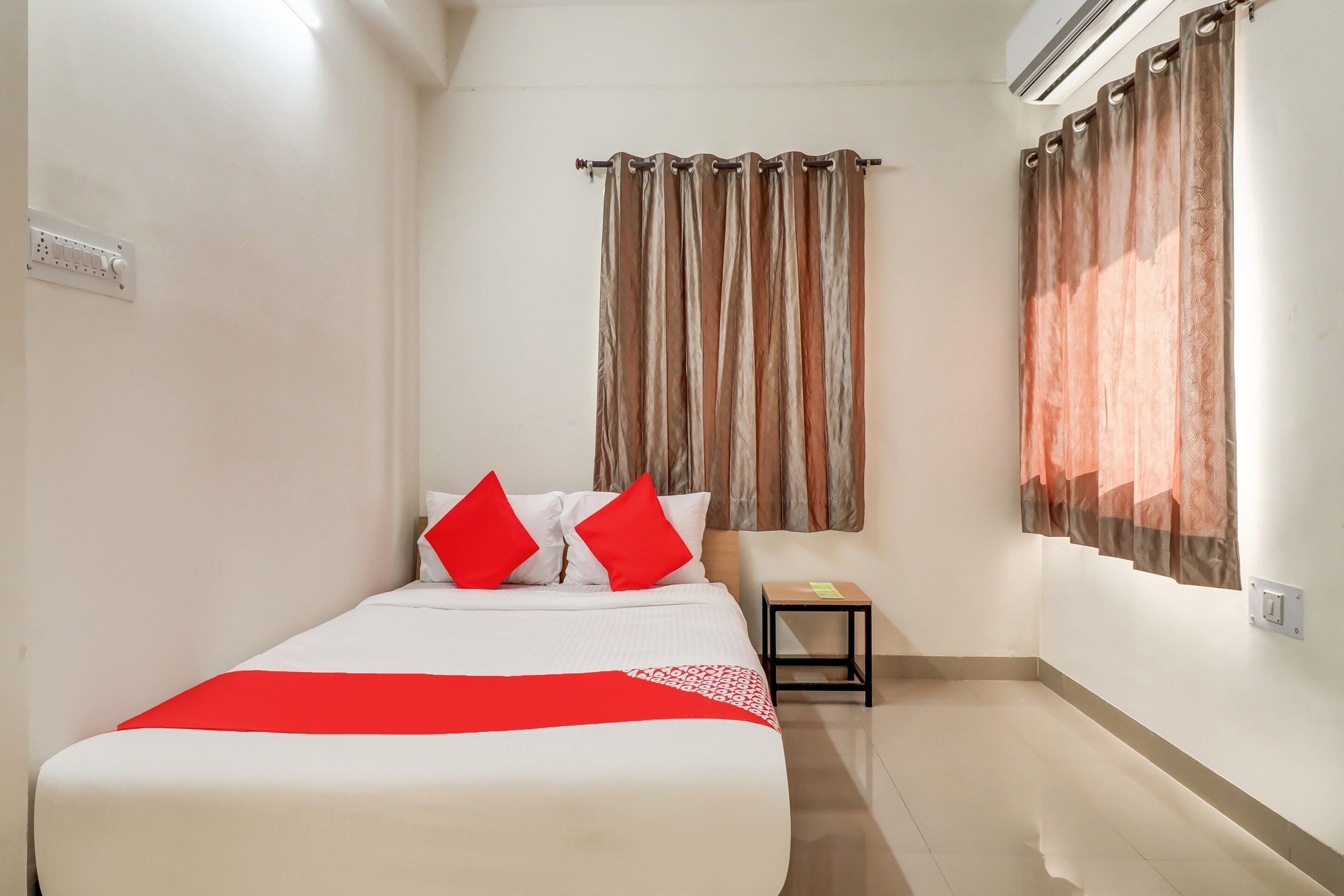 A hotel room with a bed, two red pillows, a small table, and a window with curtains.
