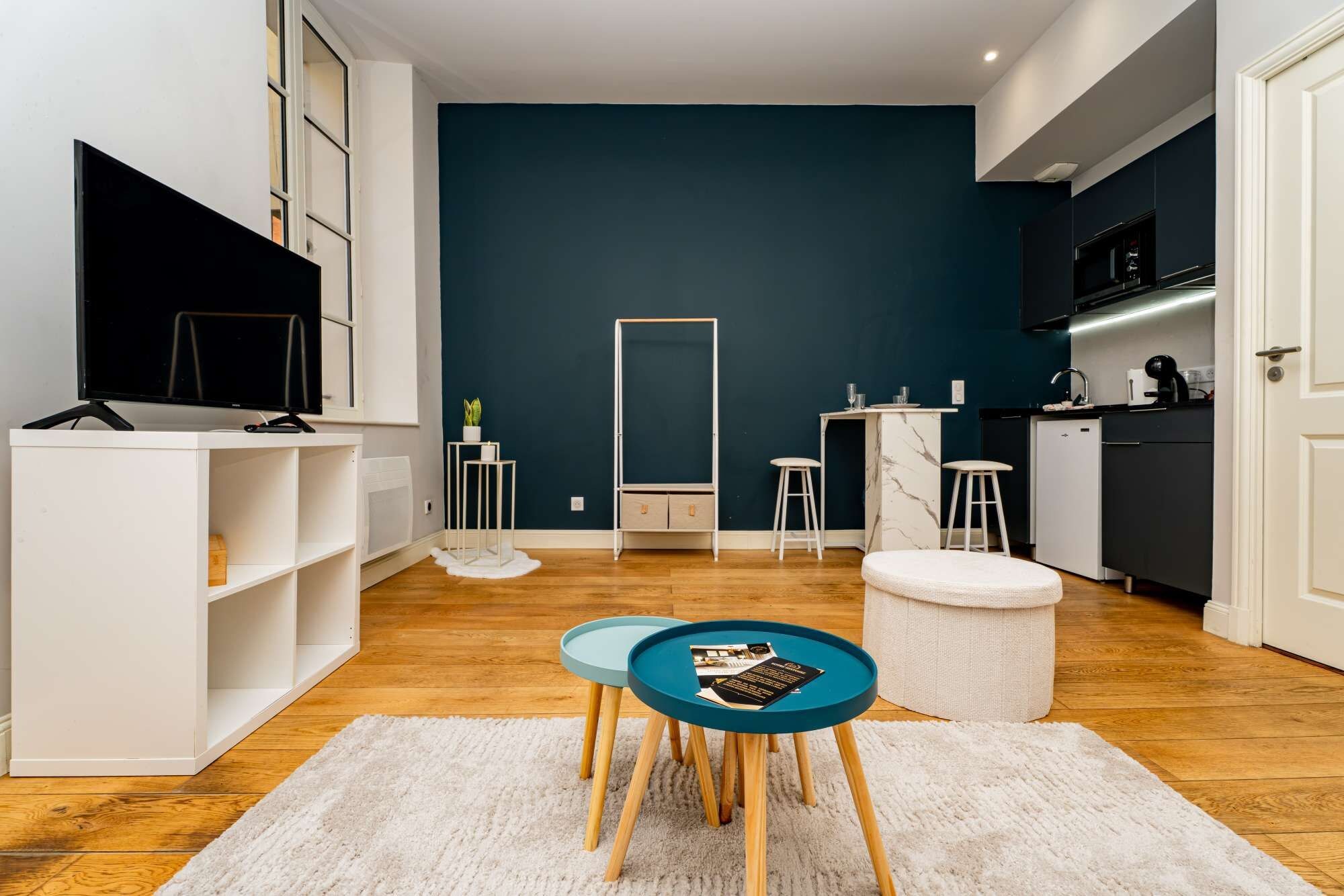 Un espacio habitable moderno con una pared azul oscuro de acento, un mueble blanco para entretenimiento, un área de cocina con microondas y una zona de estar con una mesa redonda y taburetes.